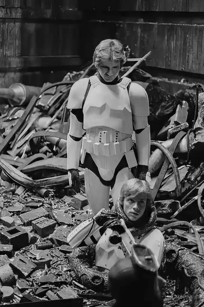 A black-and-white image shows two young boys dressed as Star Wars stormtroopers amidst rubble and debris. The taller boy…