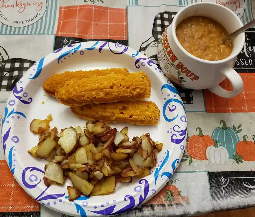 A plate of fried potatoes and two pieces of bread sits next to a mug of chicken soup on a Thanksgiving-themed tablecloth…