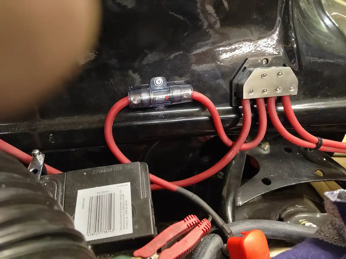 A close-up photo shows a car's engine bay with red electrical wires connected to a terminal block and a fuse holder. A black…