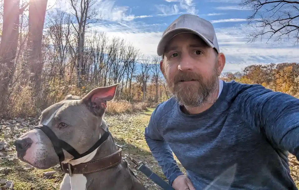 A man with a beard and cap takes a selfie outdoors with a grey and white pit bull-type dog wearing a harness and leash. The…