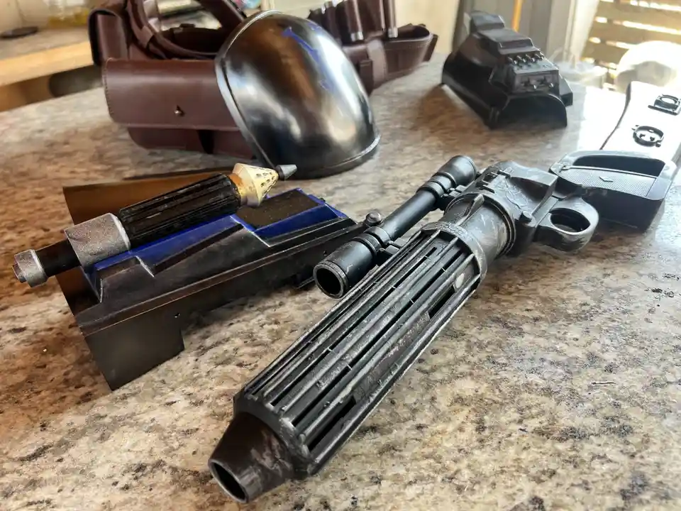 A collection of Star Wars cosplay props, including a detailed blaster rifle and a smaller blaster pistol, are laid out on a…