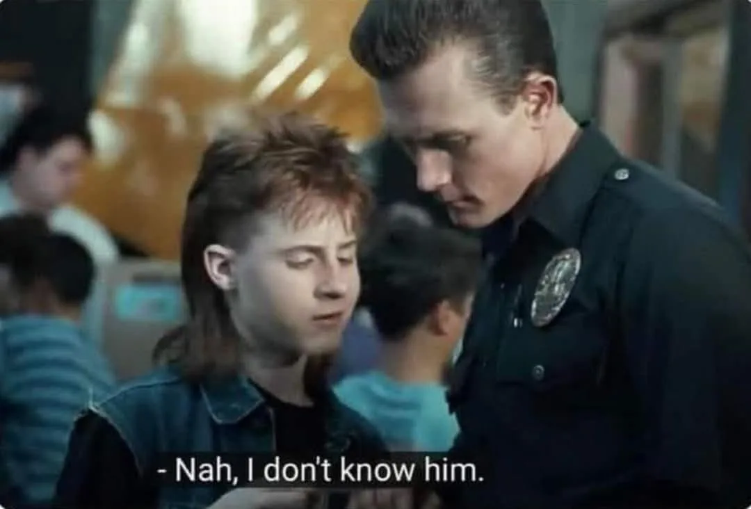 A still from a 1980s film shows a young man with a mullet and denim jacket looking down while a police officer leans in to…