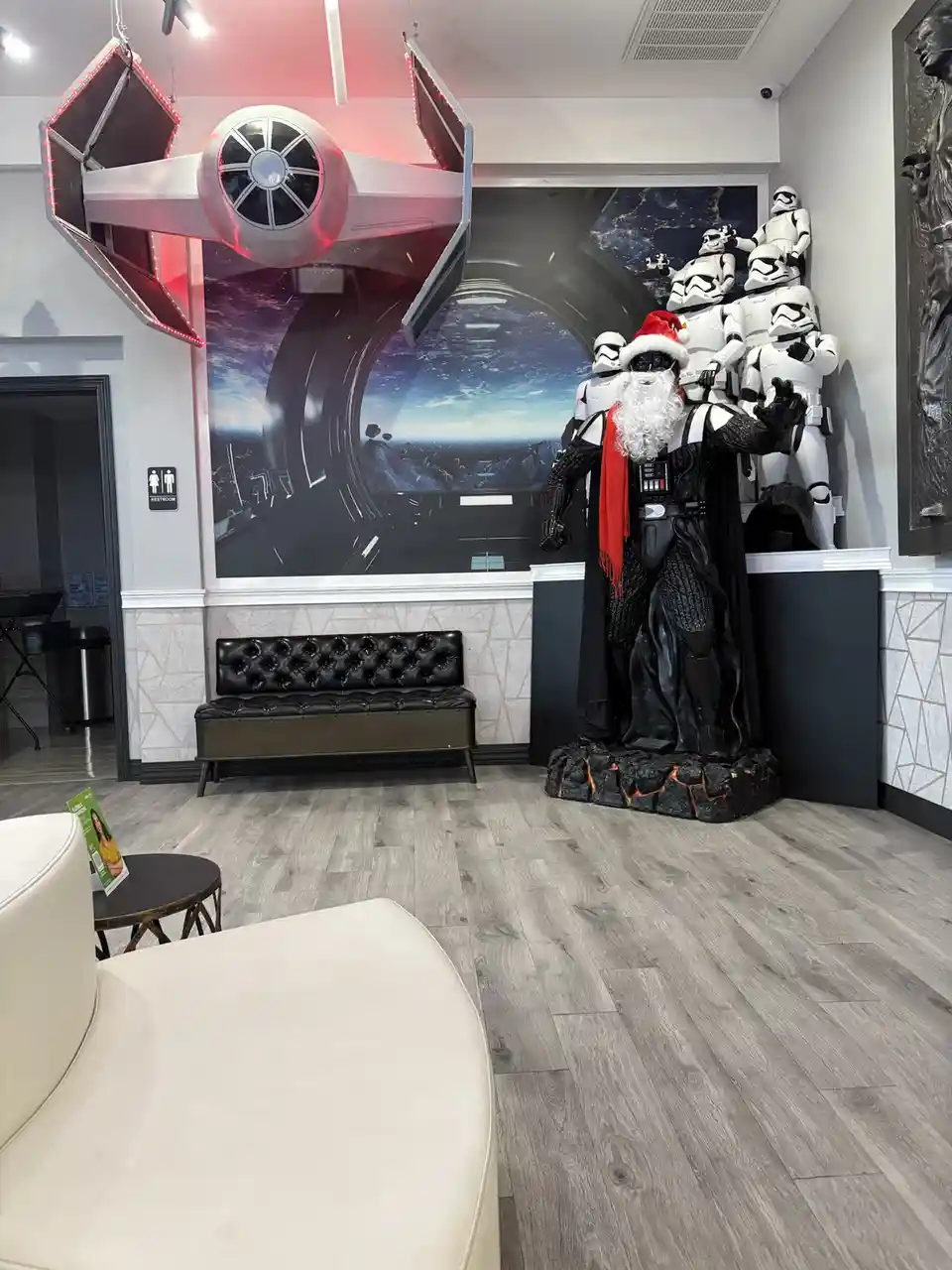 A festive indoor scene features a life-sized Darth Vader statue wearing a red Santa hat and a red scarf, standing on a…