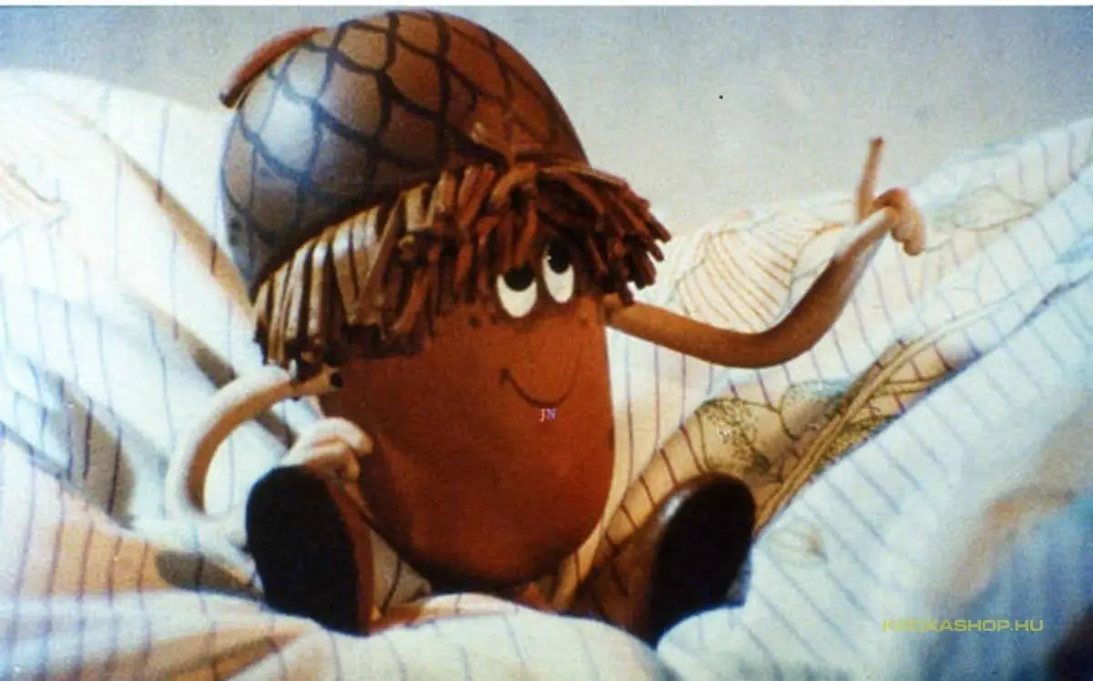 A brown, round, animated character with a netted helmet and frizzy hair sits in bed under striped sheets, smiling and…