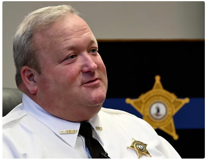 A middle-aged white man in a white sheriff's uniform with a black tie and gold sheriff's badge on his chest, looking…
