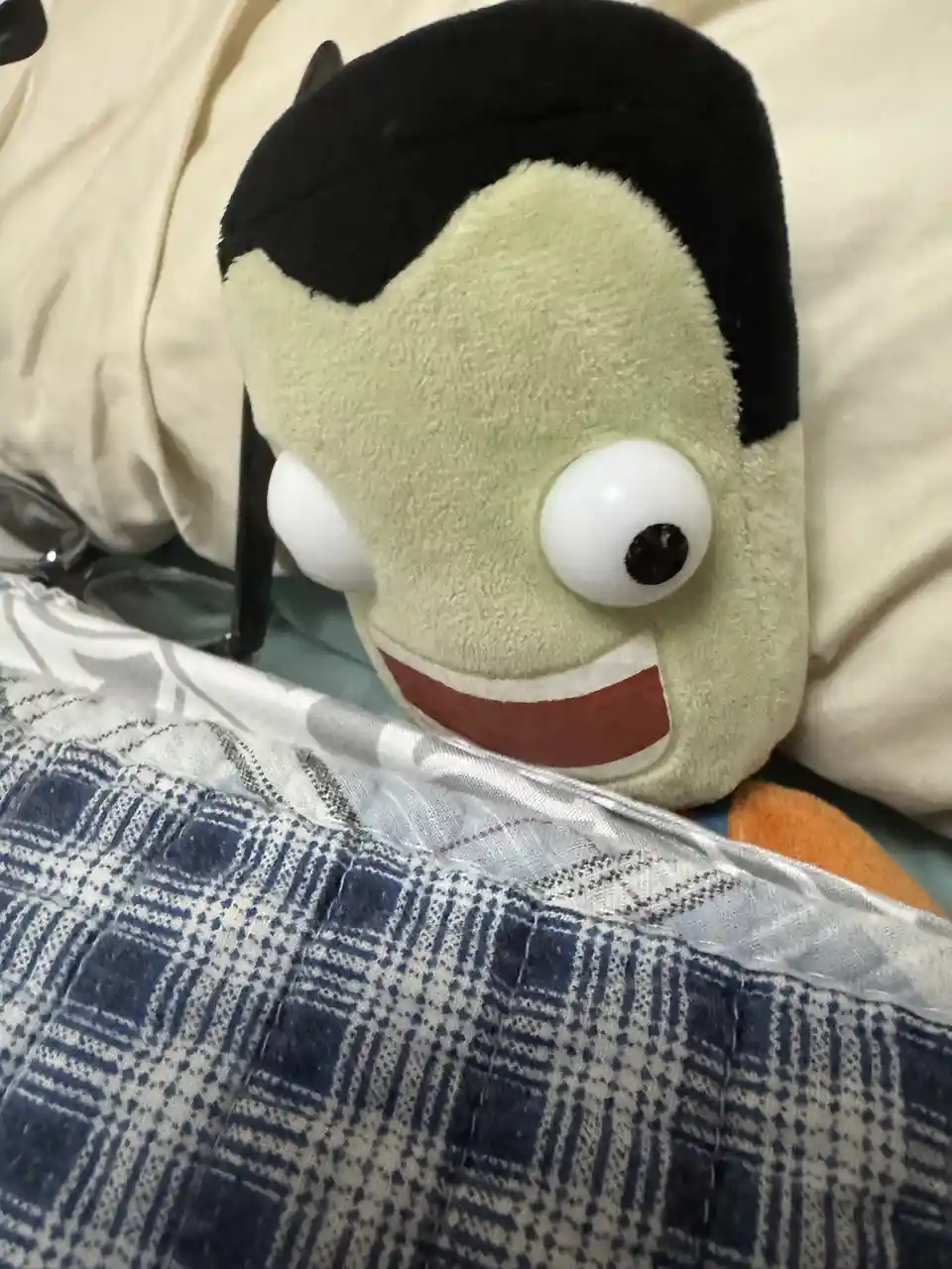A green, plush toy resembling a cartoonish Frankenstein's monster with black hair and a wide grin peeks out from under a…