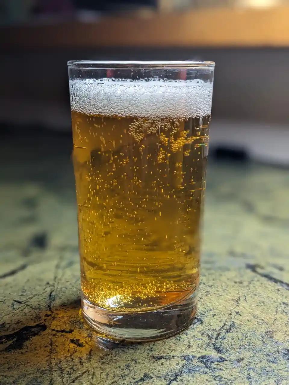 A tall glass of golden beer with a frothy white head and visible carbonation bubbles sits on a speckled countertop. The…