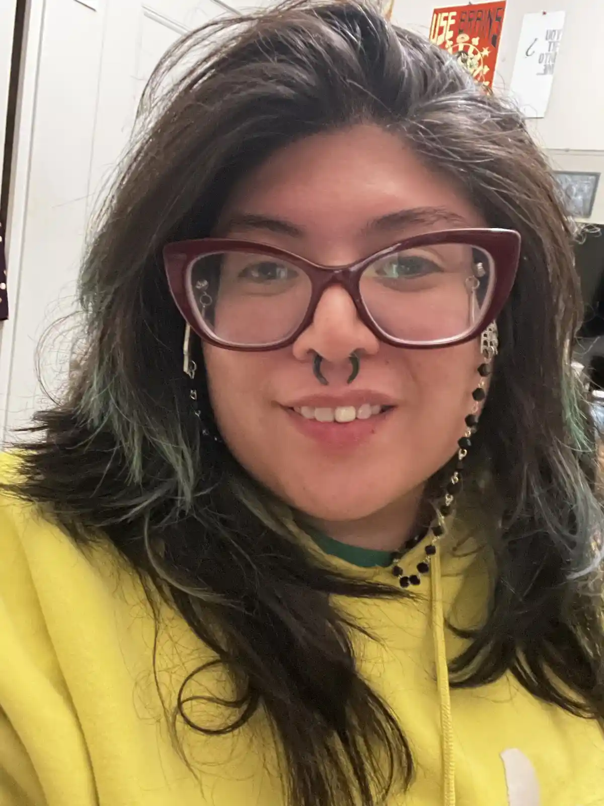 A person with dark hair and green highlights, wearing maroon cat-eye glasses, a yellow hoodie, and a nose ring, smiles at…