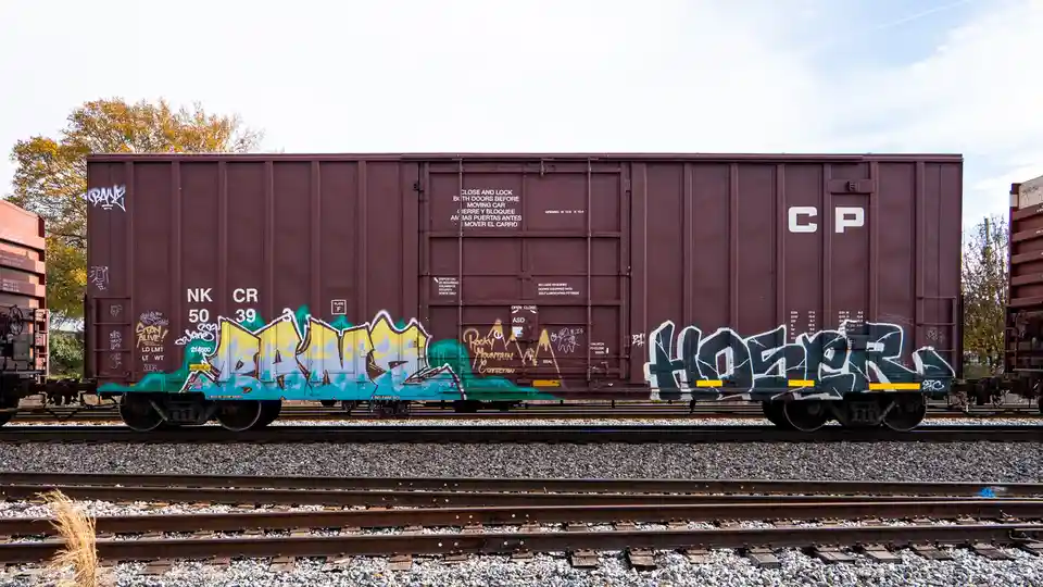 A maroon freight train car covered in colorful graffiti sits on railway tracks under a partly cloudy sky. The car has…