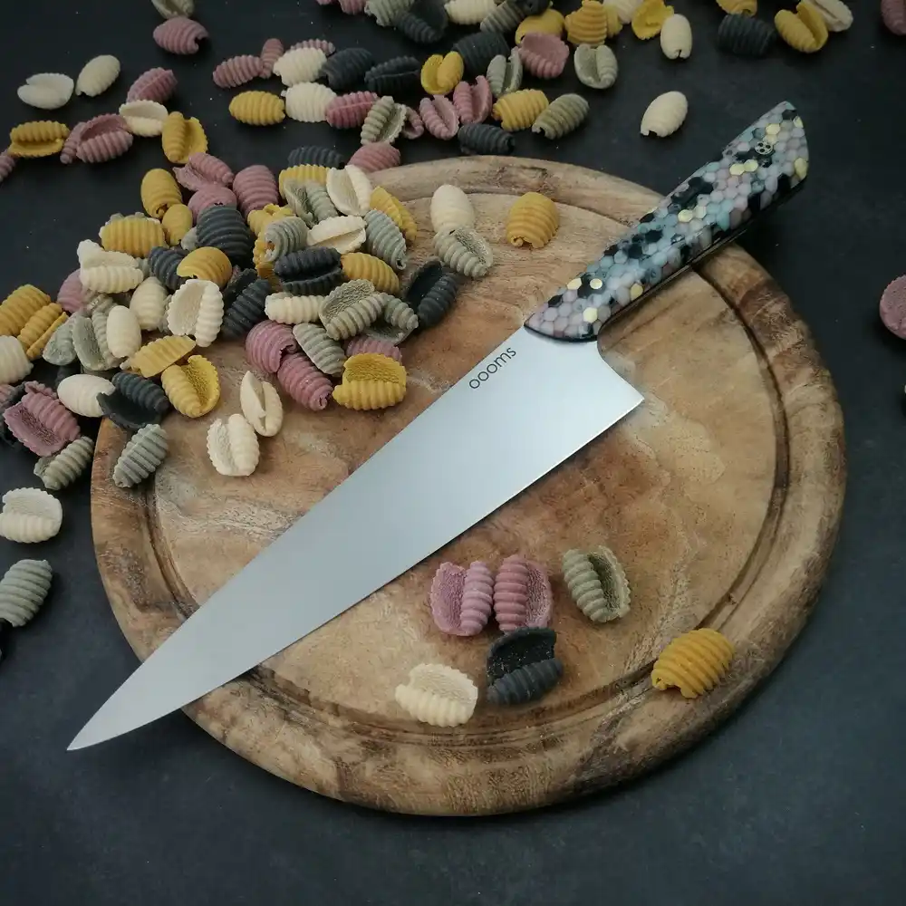 A chef's knife with a colorful, speckled handle rests on a wooden cutting board surrounded by multicolored, shell-shaped…