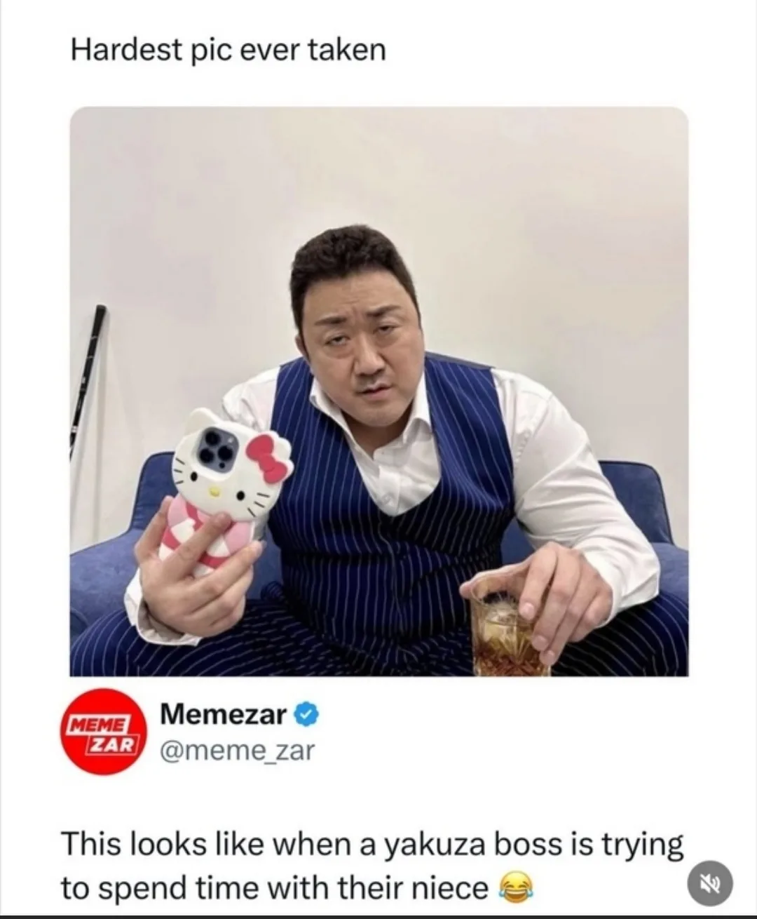 A man in a blue pinstripe vest and white shirt sits on a blue couch holding a Hello Kitty phone case and a glass of whiskey…