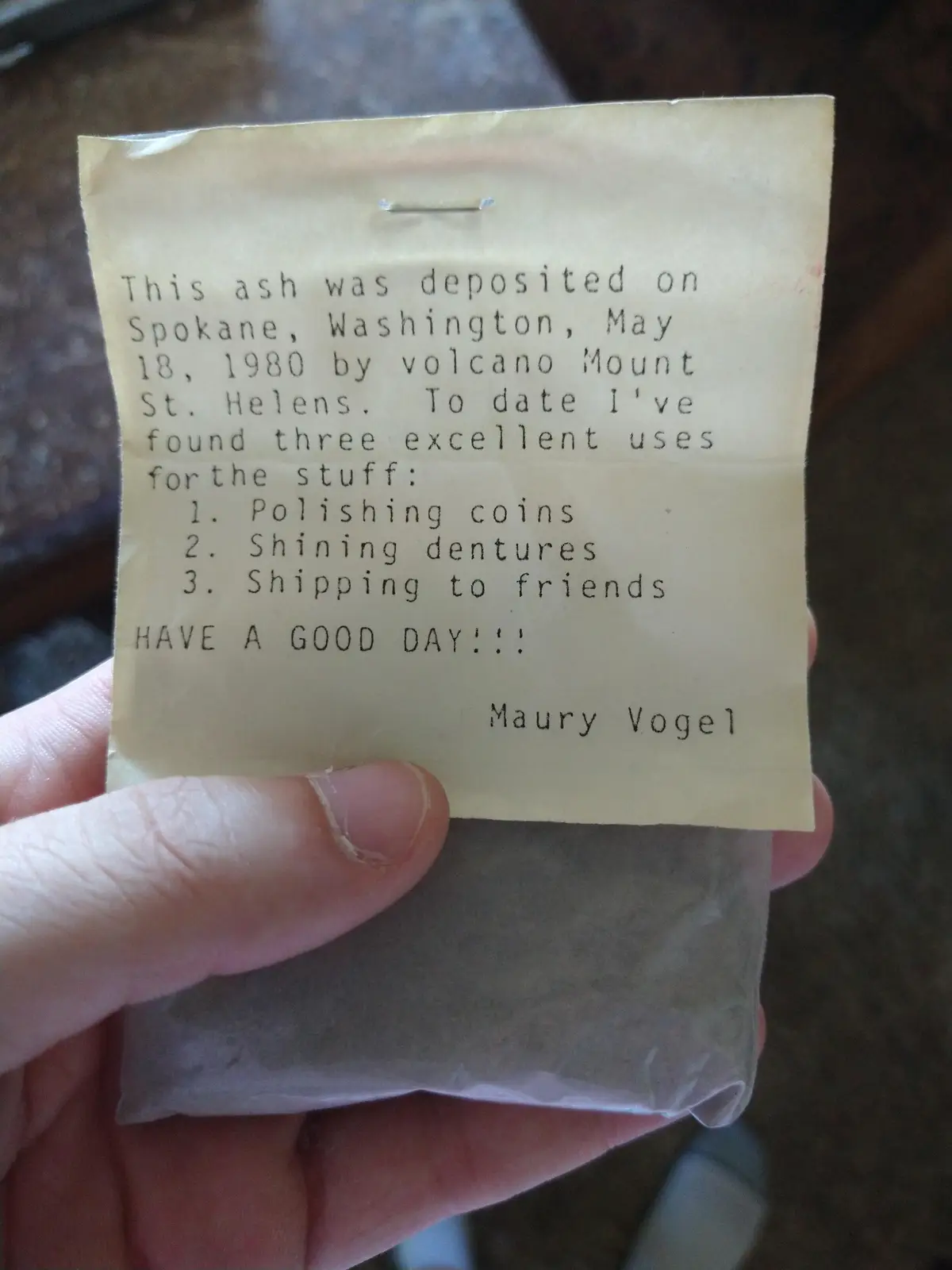 A hand holds a small plastic bag containing volcanic ash, with a typed note attached. The note, signed by Maury Vogel…