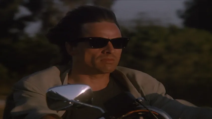a man with dark sunglasses and slicked-back hair rides a motorcycle, looking intensely forward. The overlaid text reads…