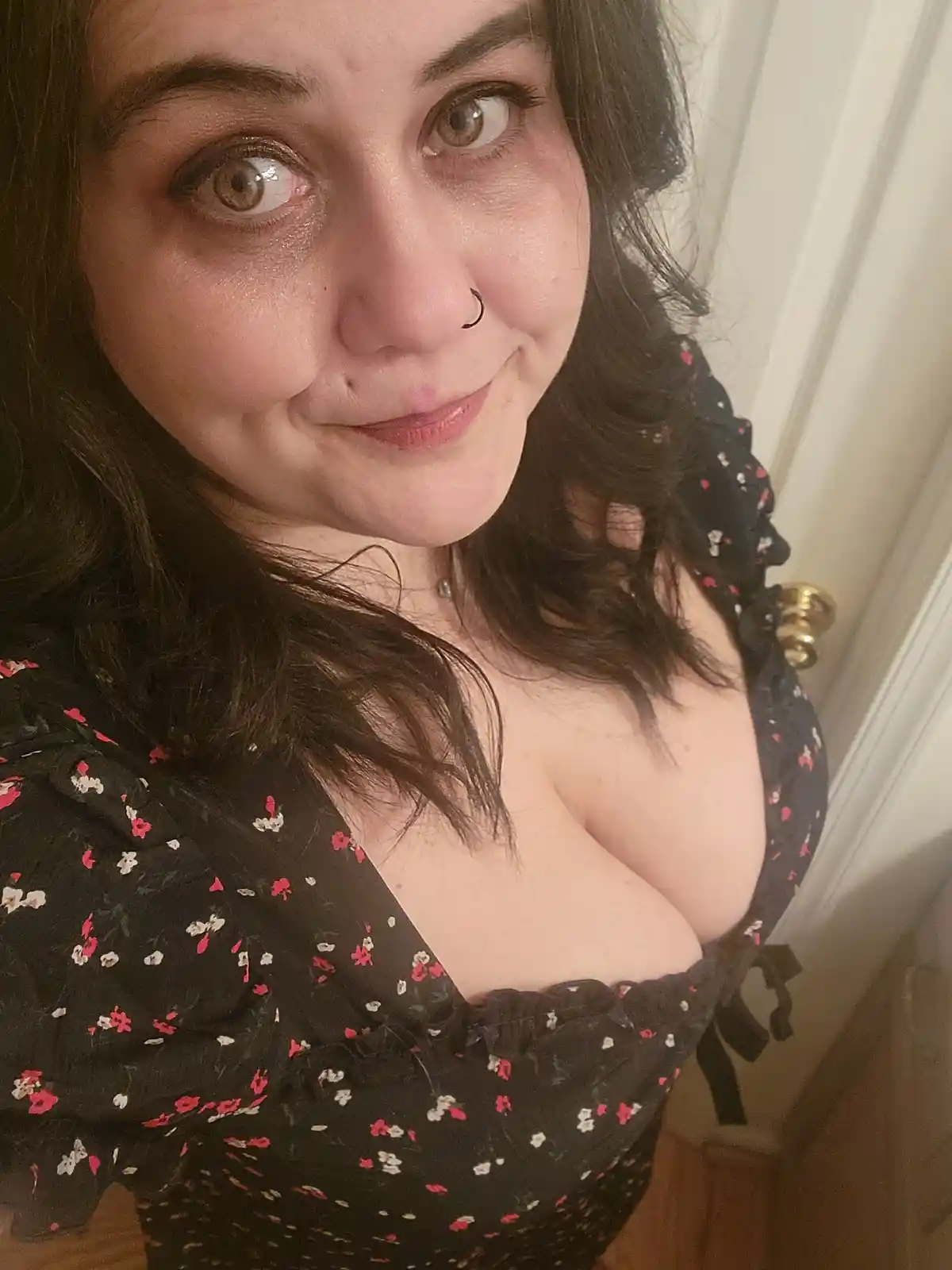 A woman with dark hair and a nose ring looks at the camera with wide eyes and a slight smirk, wearing a black floral top…