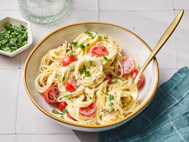 A bowl of creamy pasta with halved cherry tomatoes and chopped parsley is shown with a gold fork resting in it. A small dish…