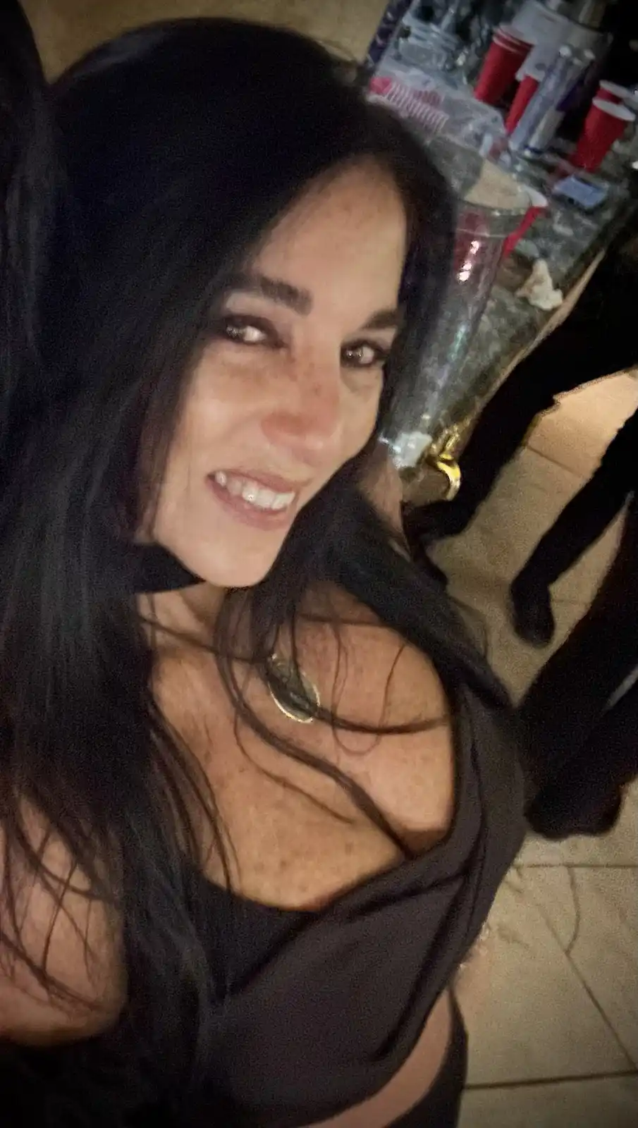 A woman with long dark hair and a black top smiles at the camera in a dimly lit, crowded setting with red plastic cups and…