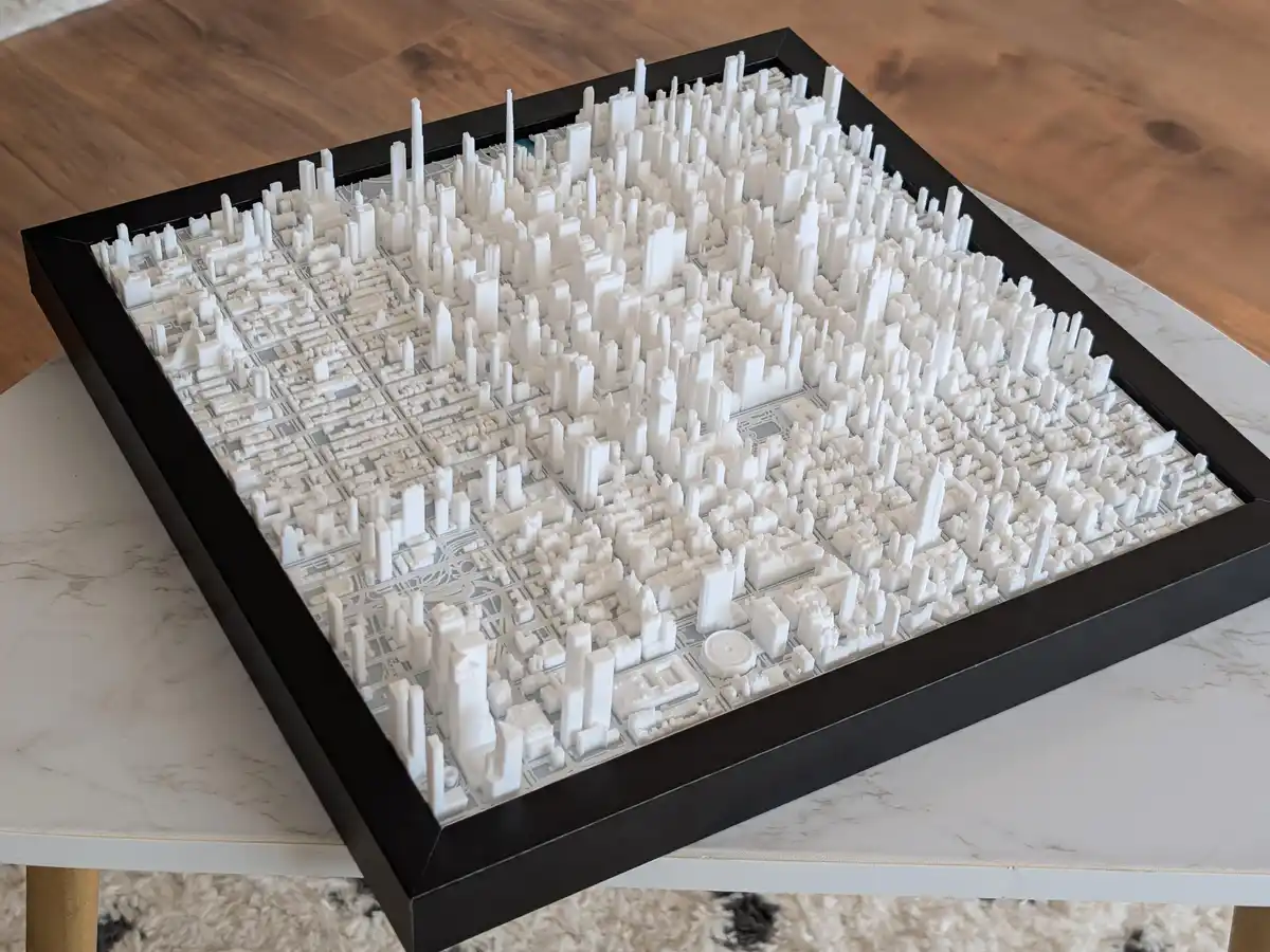 A 3d-printed white cityscape model in a black frame sits on a white marble table, showing dense buildings and streets. The…