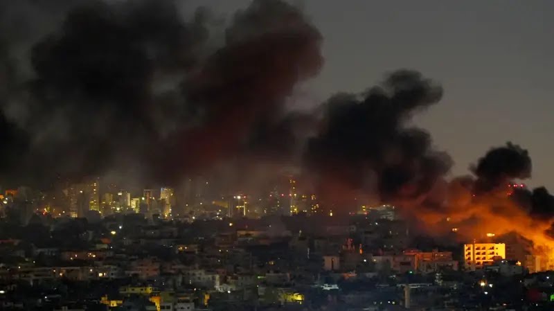 A cityscape at dusk or night with large plumes of black smoke rising from multiple fires, illuminating the sky with orange…