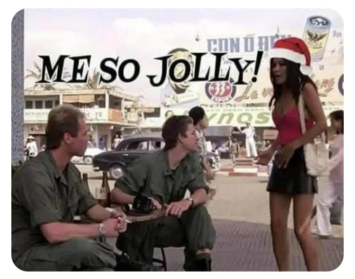 Two men in military-style uniforms sit on stools outdoors, looking up at a woman in a red Santa hat and pink top. The text…