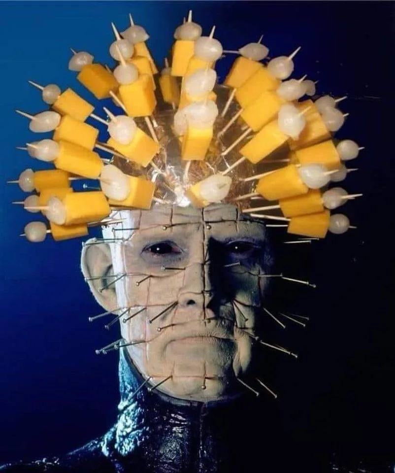 A close-up of the horror character Pinhead, whose face is covered in metal pins, with a crown made of cheese cubes and…