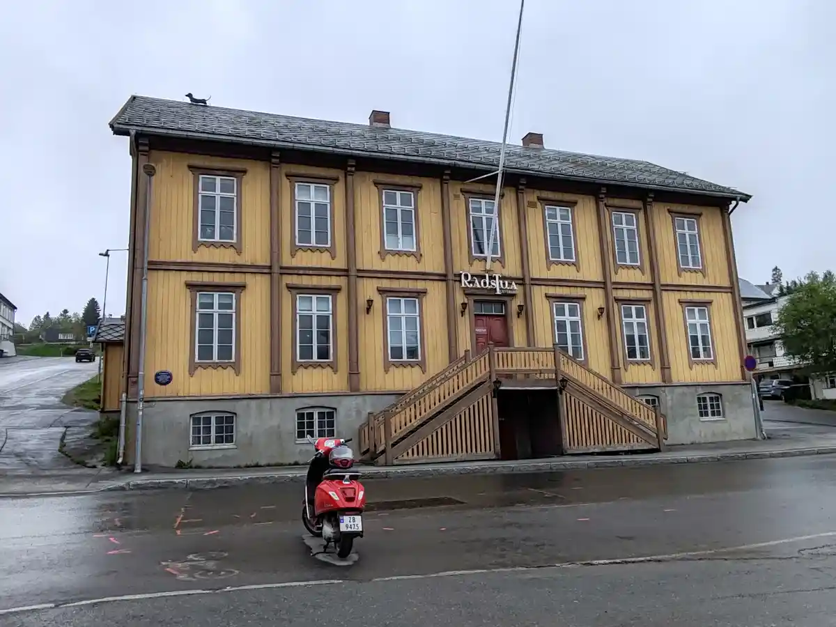 A yellow wooden building with a red door and stairs, a red scooter parked in front, and a small black dog figurine on the…