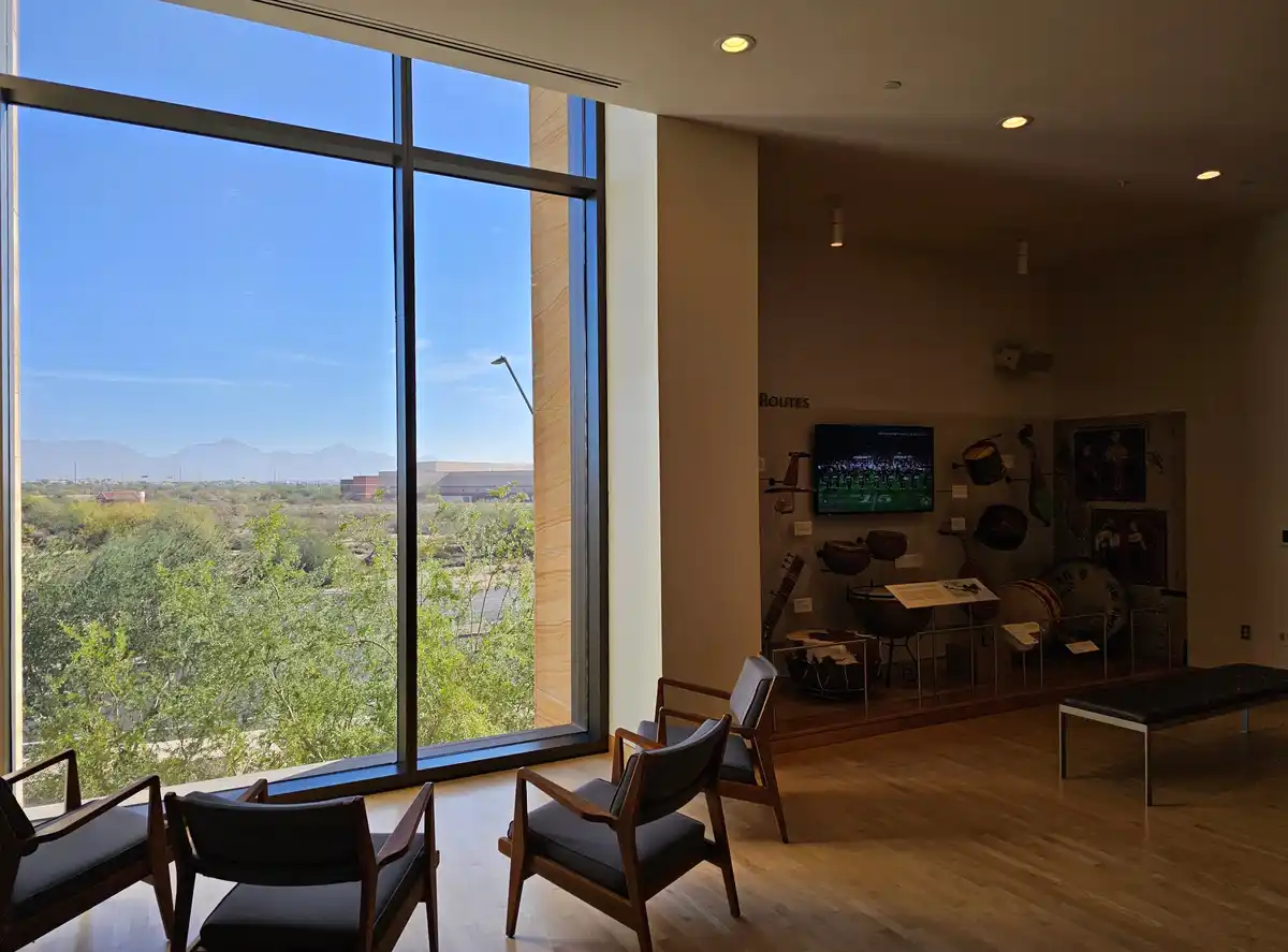 A museum room with large windows overlooking a desert landscape and distant mountains, featuring wooden chairs and a display…