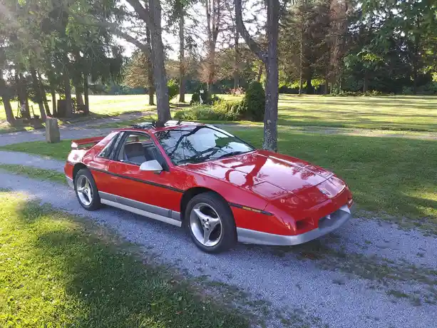 A red Pontiac Fiero sports car is parked on a gravel driveway surrounded by grass and trees. The car has a black stripe…