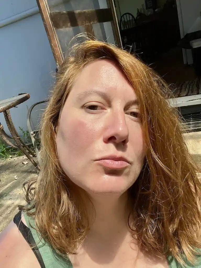 A close-up selfie of a woman with reddish-brown hair and a green tank top, looking directly at the camera with a slightly…