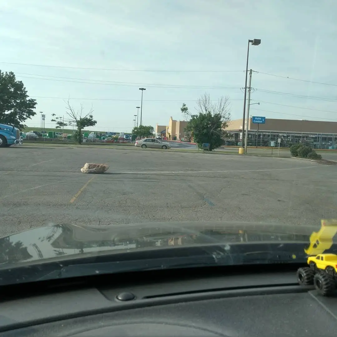 A photo taken from inside a car shows a large rock sitting in the middle of a parking lot, blocking a parking space. The…