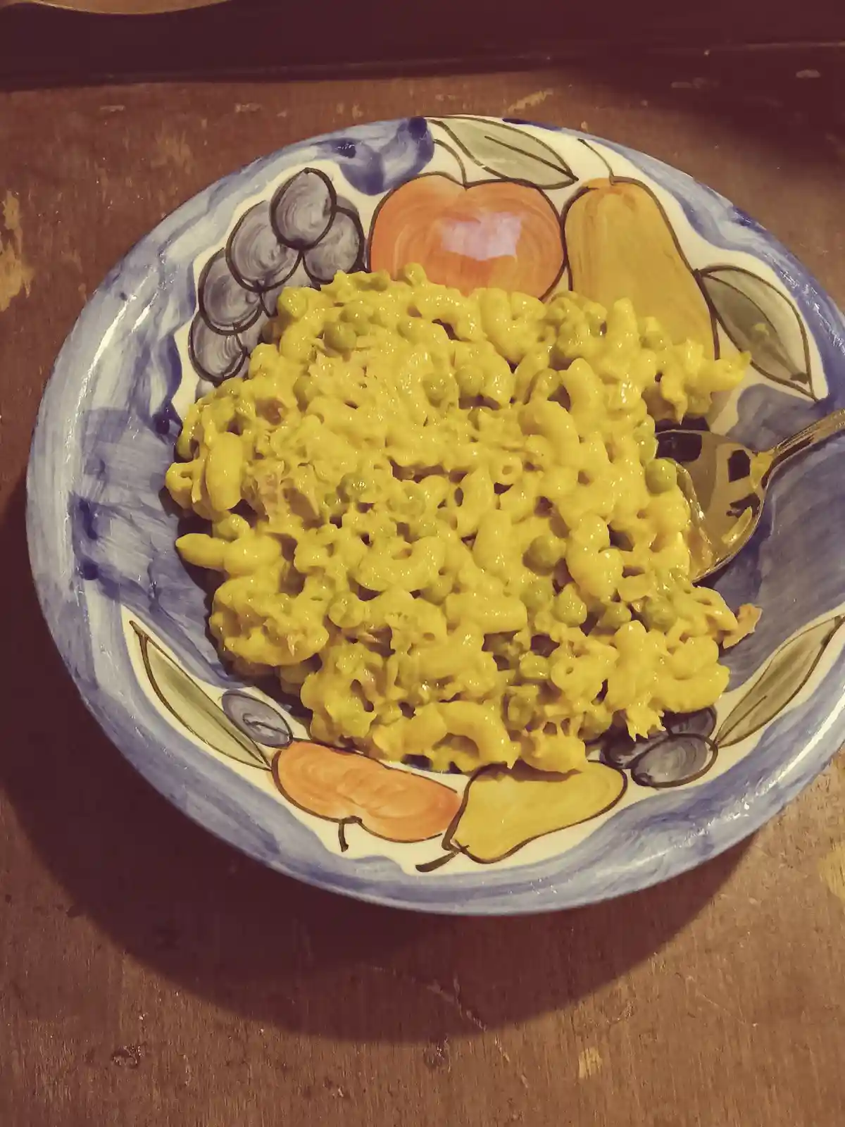 A bowl of macaroni and cheese mixed with green peas sits on a decorative plate with painted fruit patterns, resting on a…