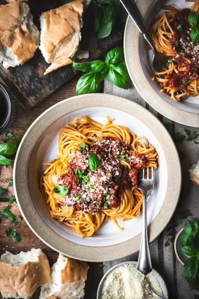 An overhead shot of two bowls of spaghetti with meat sauce, fresh basil, and grated cheese, surrounded by torn bread and…