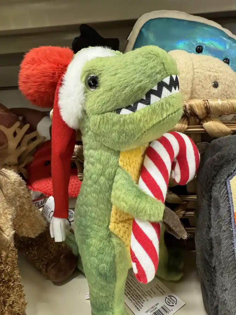 A green plush T-rex toy wearing a red and white Santa hat and holding a red and white striped candy cane, standing among…