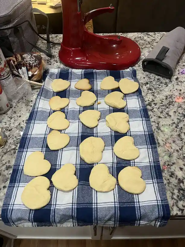 A kitchen counter displays a blue and white plaid towel with numerous heart-shaped cookies arranged on it. Some cookies are…