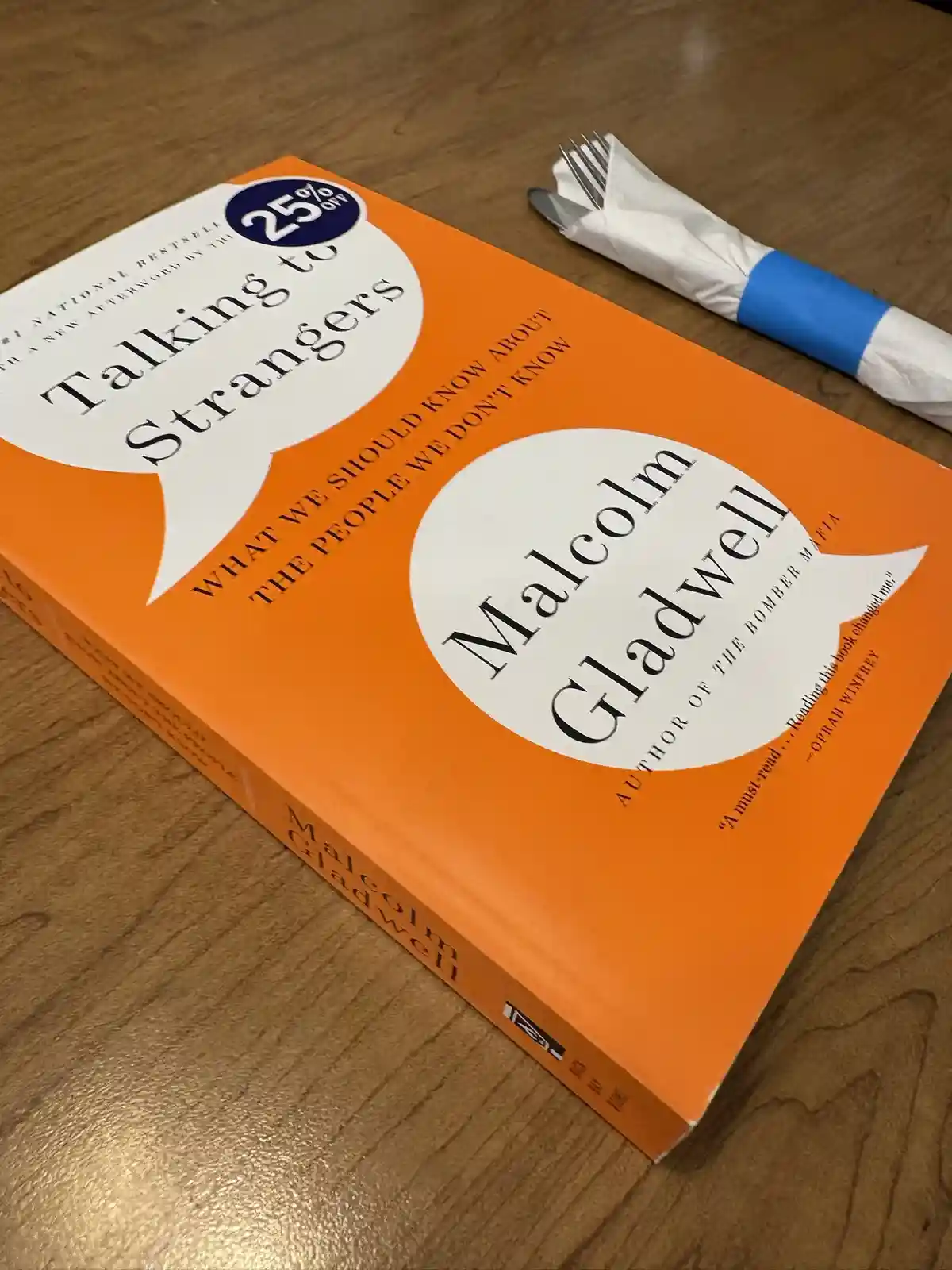 A bright orange copy of Malcolm Gladwell's book 'Talking to Strangers' lies on a wooden table, with a wrapped fork and knife…