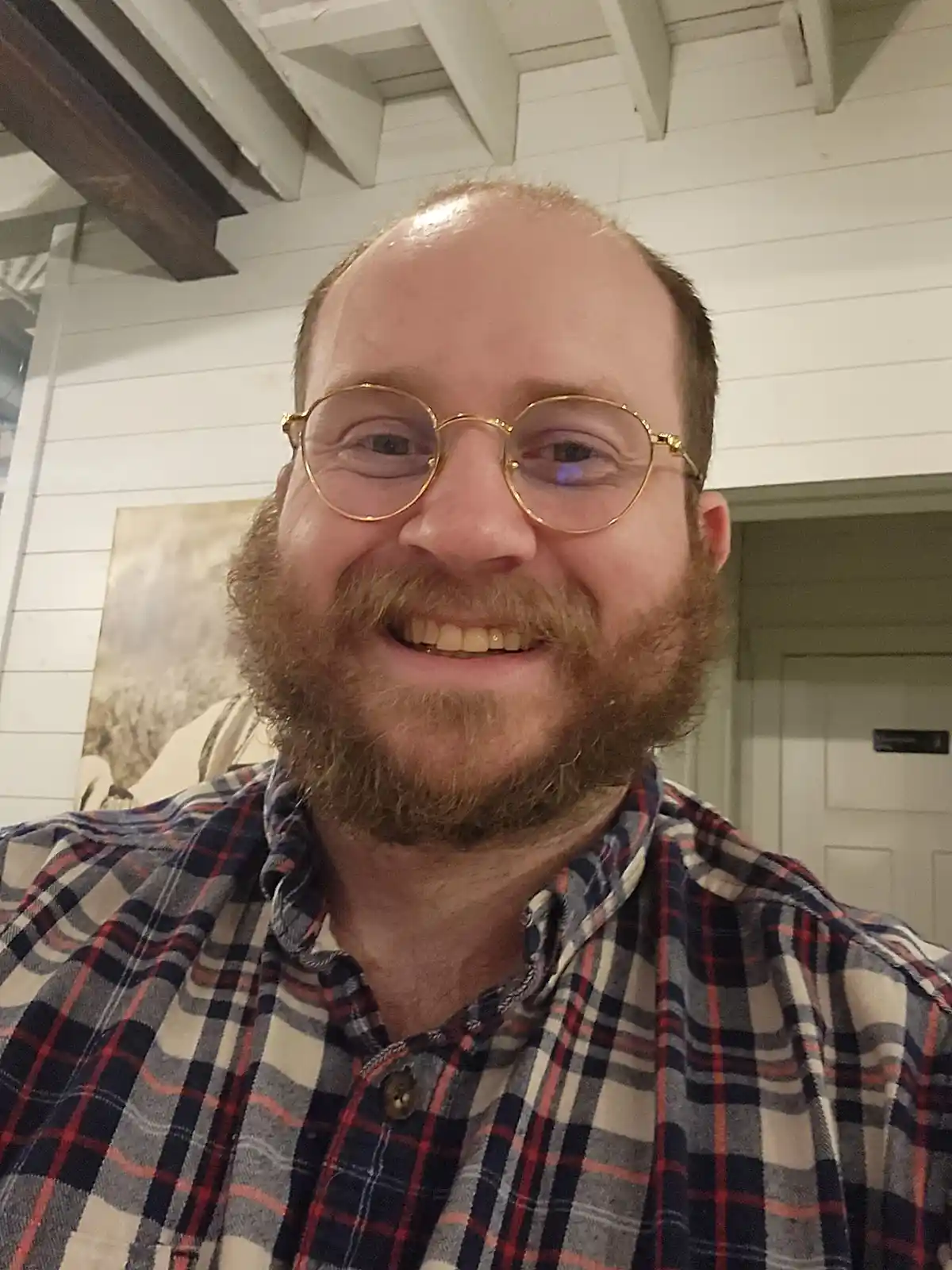 A man with a reddish beard and gold-rimmed glasses smiles at the camera while wearing a plaid shirt. The overlaid text reads…