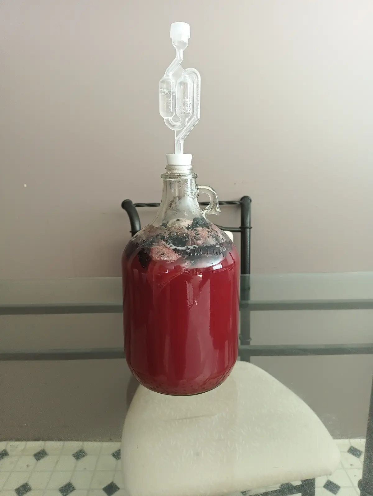 A glass carboy filled with red liquid and fruit solids sits on a white chair, topped with an airlock, suggesting homemade…
