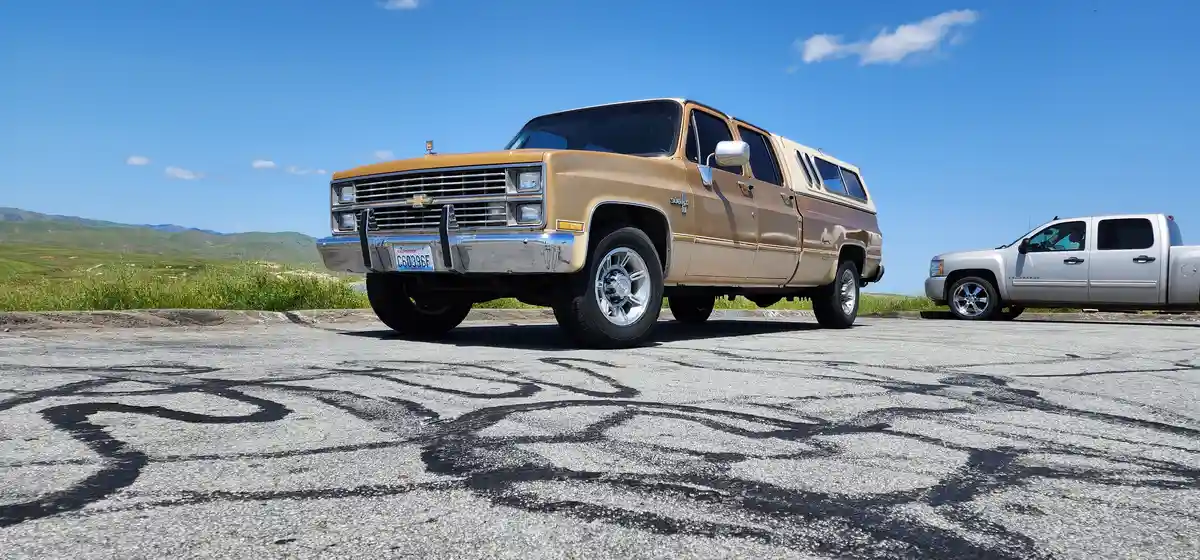 A tan 1980s Chevrolet Silverado pickup truck with a camper shell is parked next to a modern silver pickup truck on a paved…