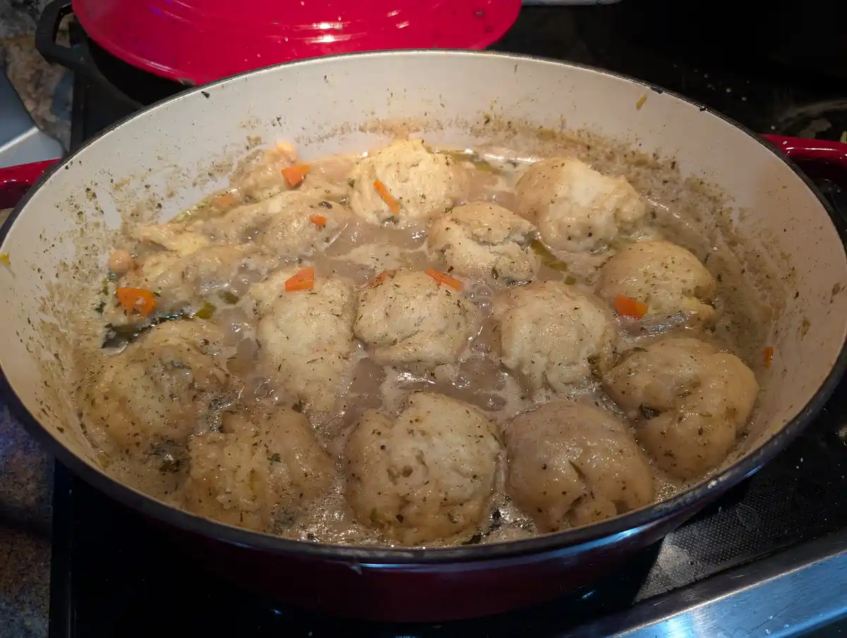 A close-up photo shows a red enameled cast iron pan on a stovetop, filled with numerous pale, lumpy dumplings simmering in a…