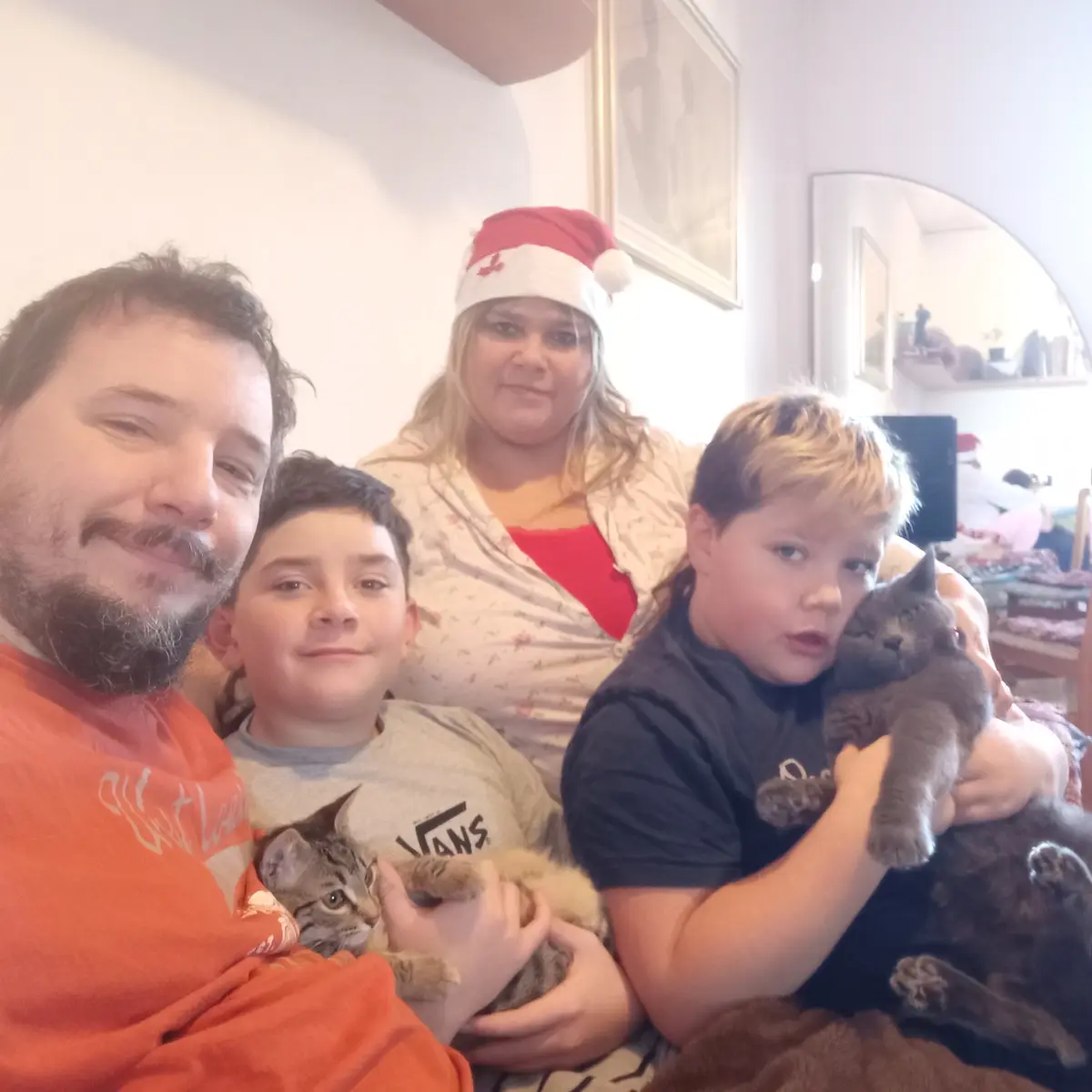 A family selfie shows four people and multiple cats in a cozy indoor setting. A woman wears a Santa hat, and a boy on the…