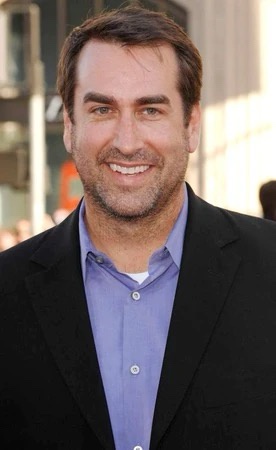 A man with dark hair and a beard, wearing a black suit jacket over a blue button-up shirt, smiles broadly at the camera. The…