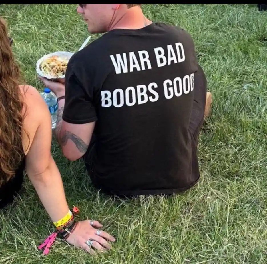 A man sitting on grass with his back to the camera wears a black t-shirt with white text reading 'WAR BAD BOOBS GOOD'. A…