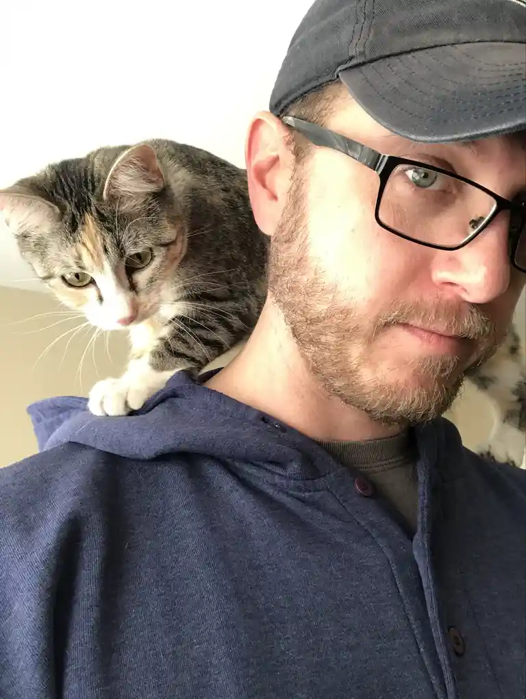 A man wearing glasses and a cap looks at the camera while a tabby cat with white and orange markings sits on his shoulder…