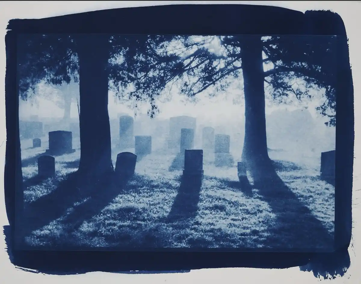 A blue-toned, vintage-style photograph of a misty cemetery with numerous tombstones and large trees casting long shadows…