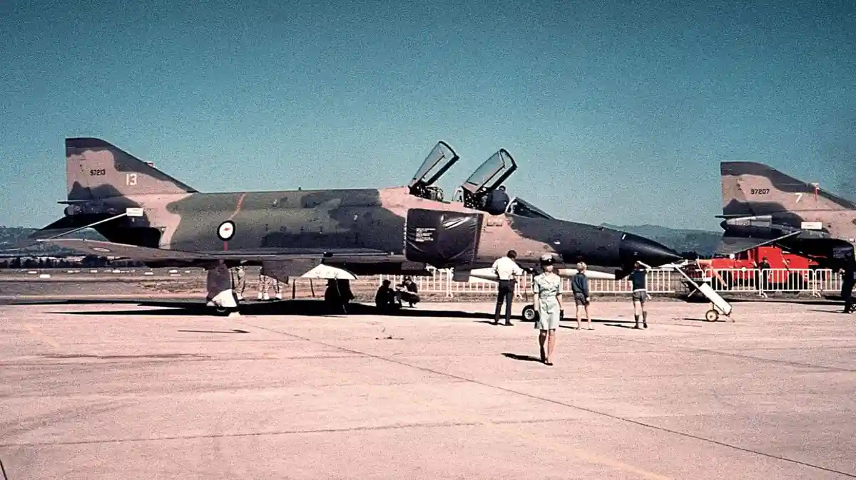 A vintage color photograph shows two military fighter jets parked on an airfield tarmac under a clear blue sky. Several…