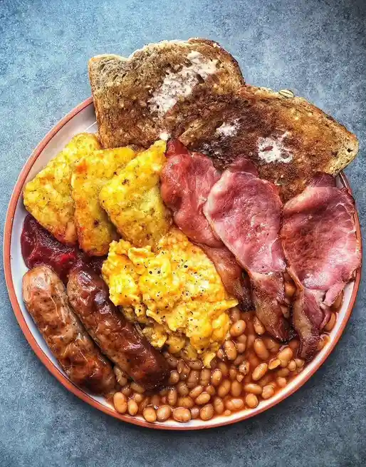 A plate of a full English breakfast featuring scrambled eggs, baked beans, sausages, hash browns, bacon, and buttered toast…