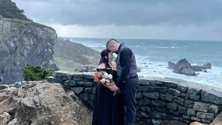 A couple stands on a rocky cliffside overlooking a stormy ocean, kissing. The woman wears a black dress and holds a bouquet…