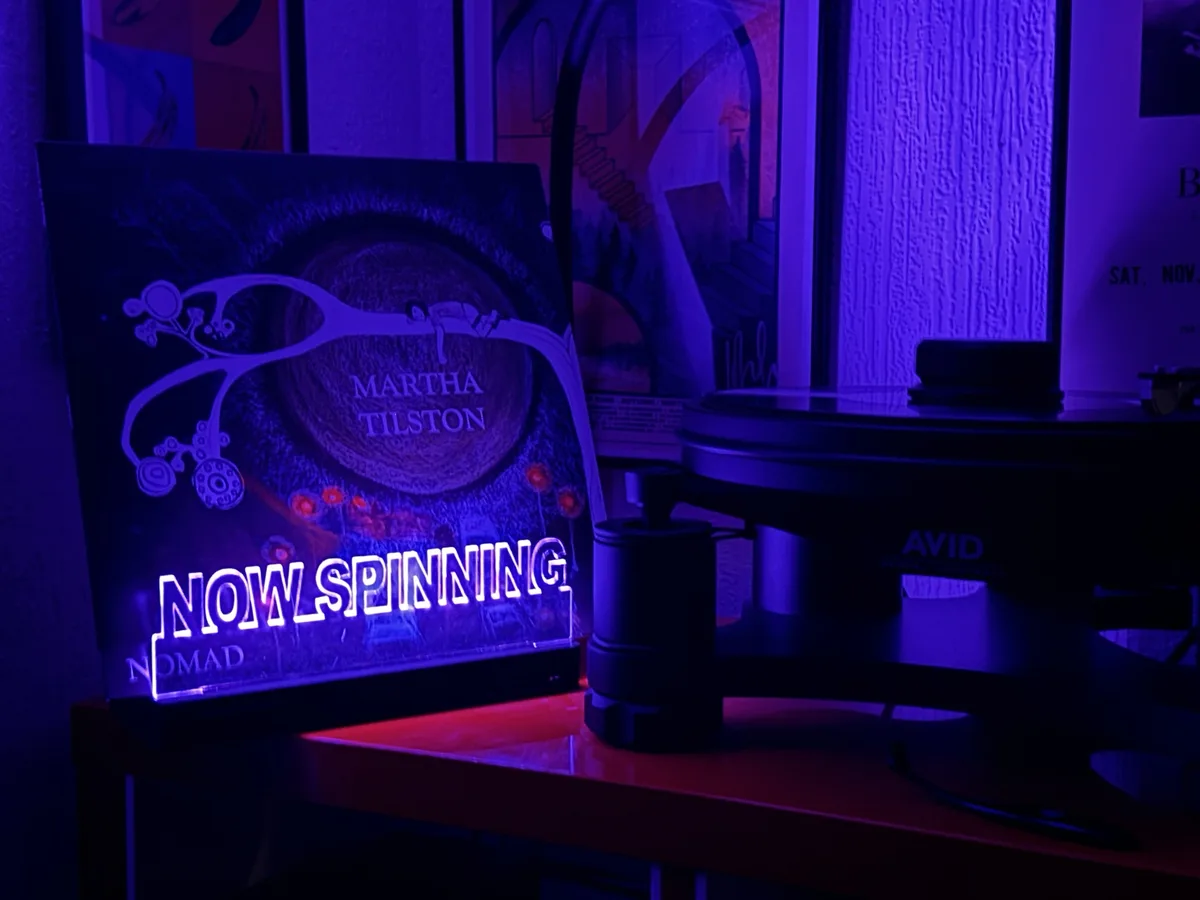 A vinyl record player sits next to a glowing 'NOW SPINNING' sign displaying the album 'Nomad' by Martha Tilston. The scene…