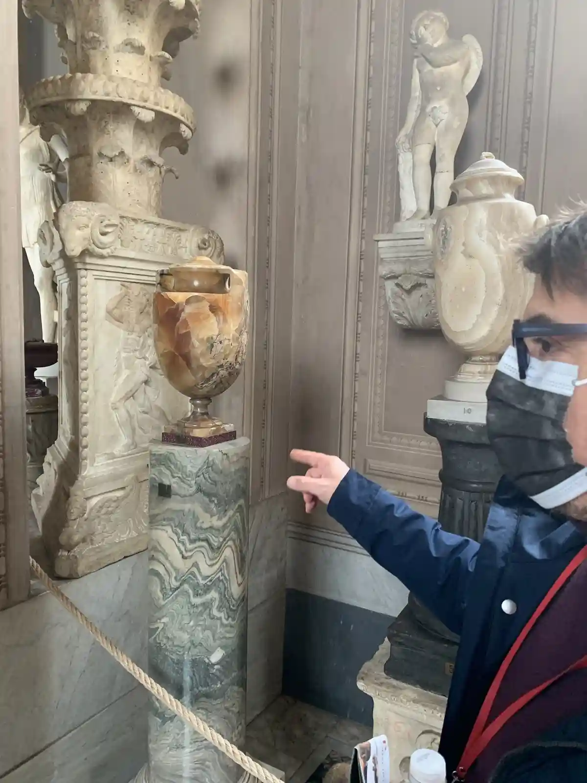 A person wearing a face mask and glasses points at a large, ornate, translucent orange-brown vase on a marble pedestal in a…