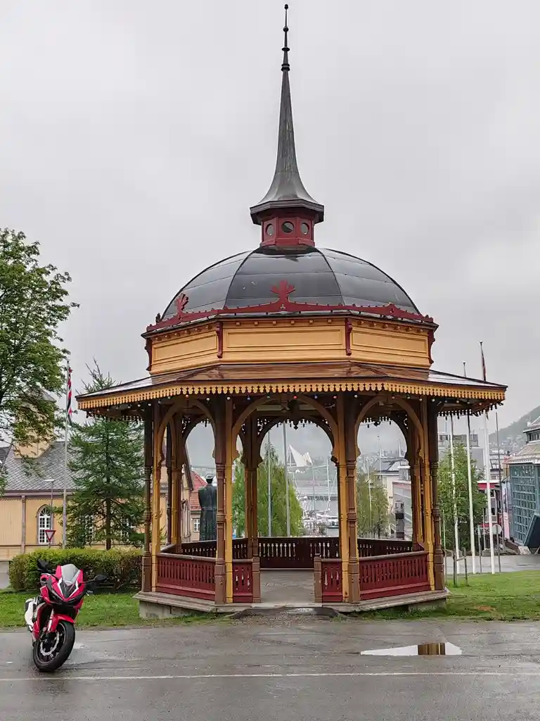 A red and yellow ornate gazebo with a black dome and spire stands in a park under an overcast sky, with a red sport…
