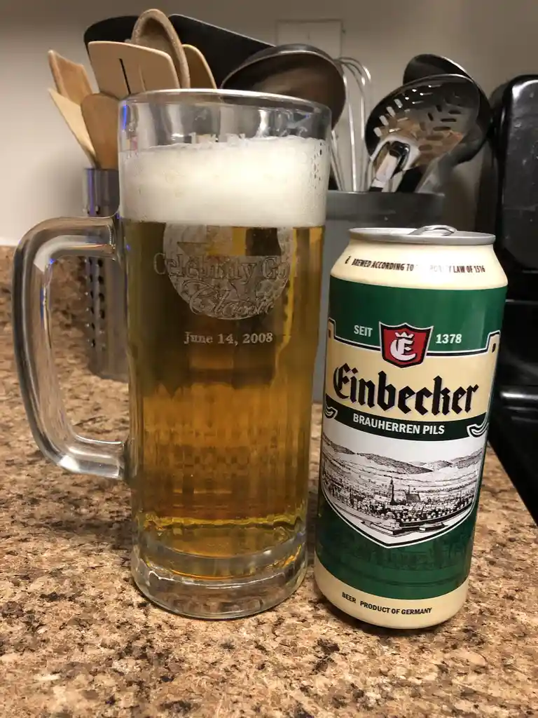 A glass mug filled with beer and topped with foam sits next to a can of Einbecker Brauherren Pils on a kitchen counter. The…